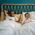 Smiling girlfriend sharing smart phone with boyfriend while lying on bed