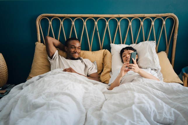 Smiling girlfriend sharing smart phone with boyfriend while lying on bed