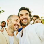 Medium shot of smiling gay couple taking selfie with friends and family after wedding ceremony on tropical beach