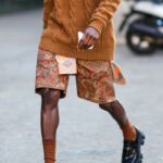 A guest wears gold large earrings, a brown half leather and half wool braided pullover, burgundy / orange / green / white print pattern shorts from Etro, brown ribbed socks, black shiny leather loafers from Prada, outside the Etro fashion show during the Milan Men's Fashion Week - Fall/Winter 2022/2023 on January 16, 2022 in Milan, Italy.