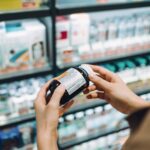 Over the shoulder view of young woman browsing through medical products and reading the label on a bottle of medicine in front of the shelves in a pharmacy