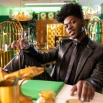 A photo of Lil Nas X in an all-black outfit, seated at a table in a room filled with gold accents.