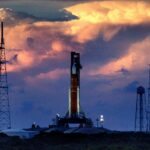 A rocket sits on the launchpad at the center of the image with sunset-lit clouds looming in the background