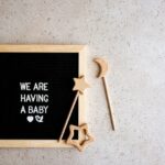 Set of baby essentials and a letter board with text “We are having a baby” on concrete background, top view. Baby announcement concept. Baby expectation. Copy space.