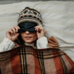 A young woman in a warm hat and sweater lifts a sleeping mask. The girl is covered with a plaid blanket and lies in bed. Close-up. Scandinavian style.