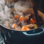 A steaming beef stew with herbs and vegetables in a blue cast iron pot with a wooden spoon on a hob stove.