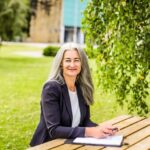 Jacqueline Carson sitting outside at a wooden table, with a clipboard in front of her, wearing a black blazer, smiling at the camera.