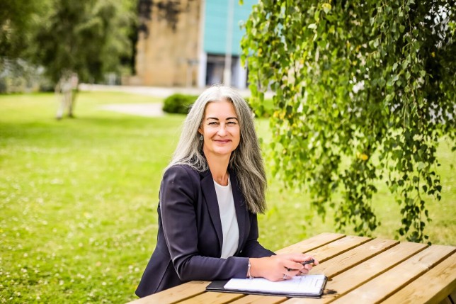 Jacqueline Carson sitting outside at a wooden table, with a clipboard in front of her, wearing a black blazer, smiling at the camera.