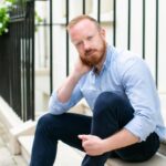 Daniel O'Shaughnessy sits on a concrete step, with ginger hair and a beard, wearing a light blue shirt and dark blue trousers, he is smiling at the camera