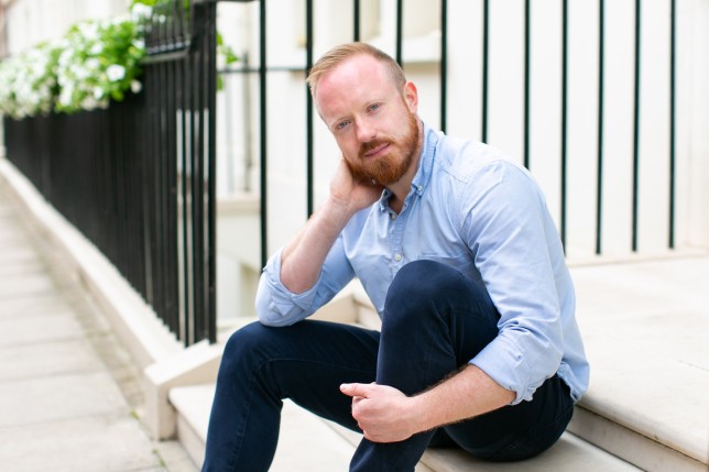 Daniel O'Shaughnessy sits on a concrete step, with ginger hair and a beard, wearing a light blue shirt and dark blue trousers, he is smiling at the camera