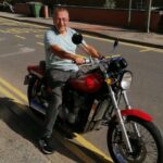 Mike Foster sitting on a red motorbike, which is stationary on a residential street.