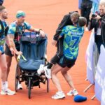 Kevin Sinfield, a man wearing bright running clothes with 'Burrow' on the back, lifts a man in a black tracksuit out of a wheelchair while two other men in the same clothes watch on