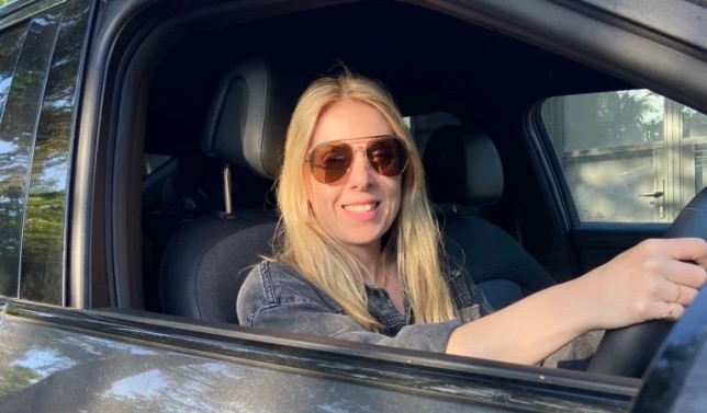 Rachel, wearing sunglasses and sitting in her car with the window rolled down and smiling to camera