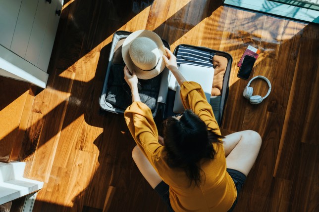 Overhead view of young Asian woman sitting on the floor in her bedroom, packing a suitcase for a trip. Getting ready for a vacation. Traveller's accessories. Travel and vacation concept