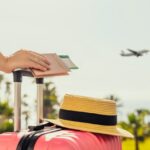 Woman with pink suitcase and passport with boarding pass standing on passengers ladder of airplane opposite sea with palm trees. Tourism concept