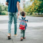 Rear view of father who leads a little boy hand in hand to kindergarten. Father and son with backpack walking in schoolyard.