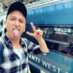 Jason Reid making peace sign and sticking tongue out next to a train. He is wearing a black cap and plaid shirt