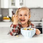 Beautiful little girl having breakfast with cereal, milk and blueberry in kitchen