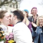 Fox Fisher, a person in a white suit, kisses their partner, who is wearing a white dress.