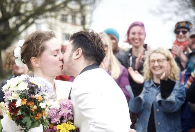 Fox Fisher, a person in a white suit, kisses their partner, who is wearing a white dress.