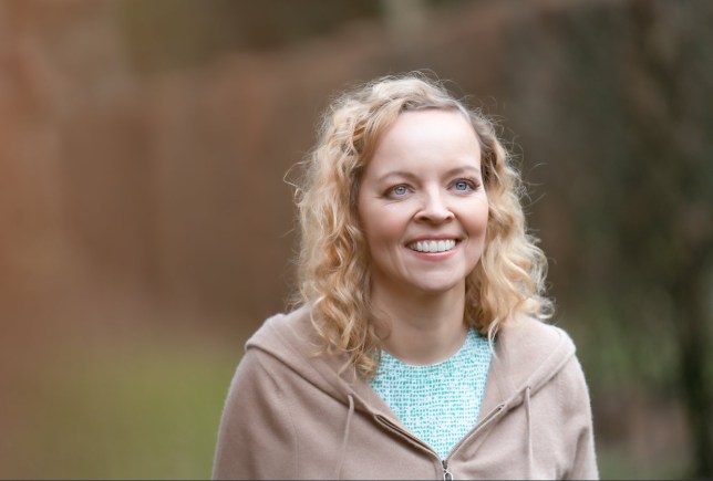 Sandra wearing a green sequinned top, with a beige hoodie on top of it. She has light blue yes and blonde hair and is smiling - the background is blurred