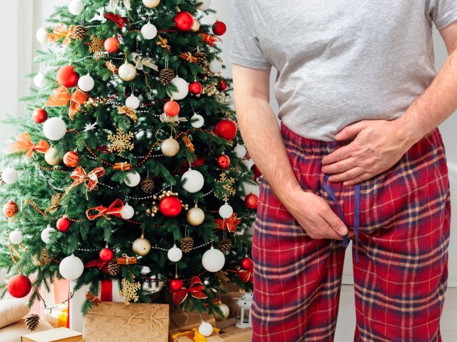 A man holding his pelvic area, while stood in front of a Christmas tree