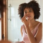 Young African woman putting cream on her face in a bathroom mirror