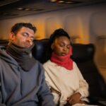 Two passengers sleeping on a flight during the night time - wearing trtl travel pillow