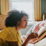 A woman lies on her stomach on a comfortable bed and reads a book.