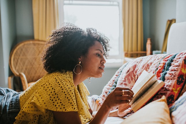 A woman lies on her stomach on a comfortable bed and reads a book.