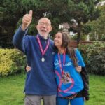 Chloe standing with her dad in the garden, their arms around each other. She's looking at him and smiling, he's looking to camera with his thumbs up