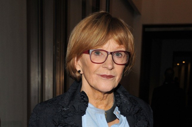 Anne Robinson wearing dark red framed glasses, with short red hair, a black jacket and blue top