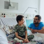 Tia sitting on her hospital bed wearing a camo t-shirt and trousers, looking at her dad who is kneeling by the side of the bed smiling back at her