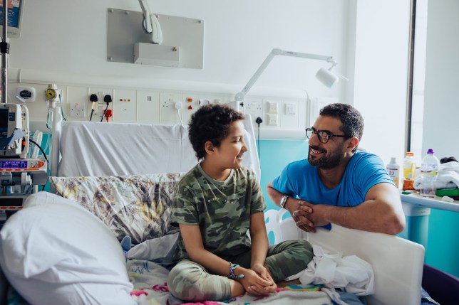 Tia sitting on her hospital bed wearing a camo t-shirt and trousers, looking at her dad who is kneeling by the side of the bed smiling back at her