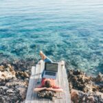 Young freelance woman works in laptop lying at backpack on wooden beach decks against Mediterranean sea