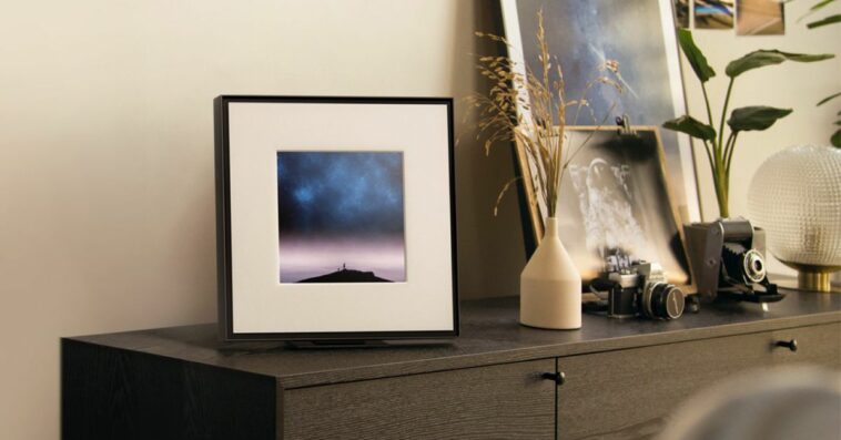 Samsung’s Music Frame speaker sitting on a brown cabinet beside a pair of cameras and some plants.