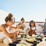 Medium wide shot of smiling female friends sharing breakfast on deck of luxury suite at tropical resort