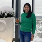 Joanne standing at a snowy viewpoint at the top of a building, holding a drink and the wearing a green jumper