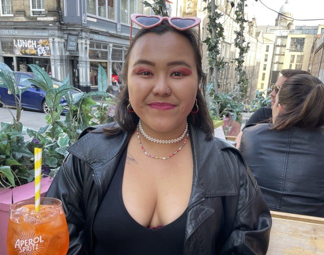 Michele sitting outside of a bar, with plants behind her - she is wearing a black jacket and top, with glasses on her head