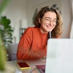 Young happy woman student using laptop watching webinar writing at home.