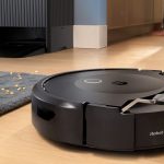 A black robot vacuum on a hardwood floor with a mop extended above it and a big, black charging dock behind it.