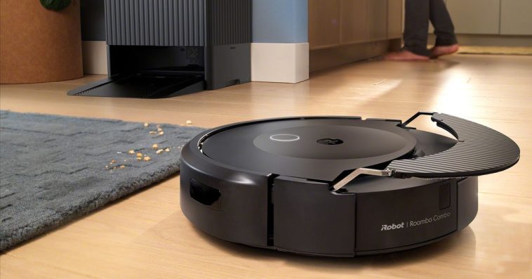 A black robot vacuum on a hardwood floor with a mop extended above it and a big, black charging dock behind it.