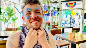 William sitting in a cafe and has glitter on their face - they are smiling happily to camera