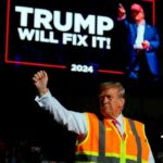 Donald Trump gestures in front of a political display board that reads ‘Trump will fix it!’