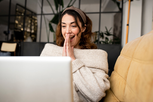 Excited woman looking at laptop screen sitting on cozy sofa at home