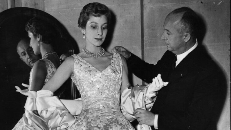 Christian Dior (1905 - 1957) French couturier, with a mannequin in one of his own designs, a satin evening gown called Blenheim.