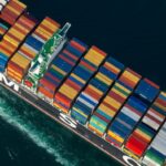 Aerial view of a tiny tugboat helping to guide a shop loaded with freight containers