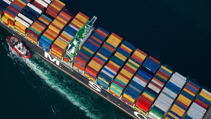 Aerial view of a tiny tugboat helping to guide a shop loaded with freight containers