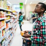 Woman standing in a supermarket aisle with a basket, looking at food products.